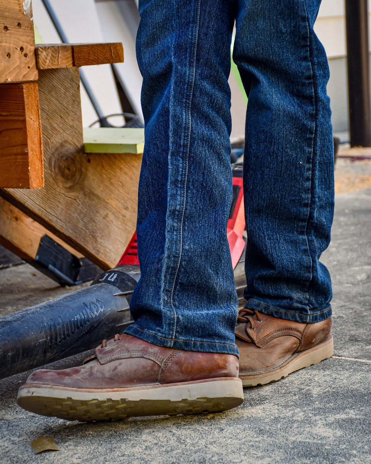 Model wearing brown leather boots and blue jeans on a job site. Stairs and leaf blower shown in background.
