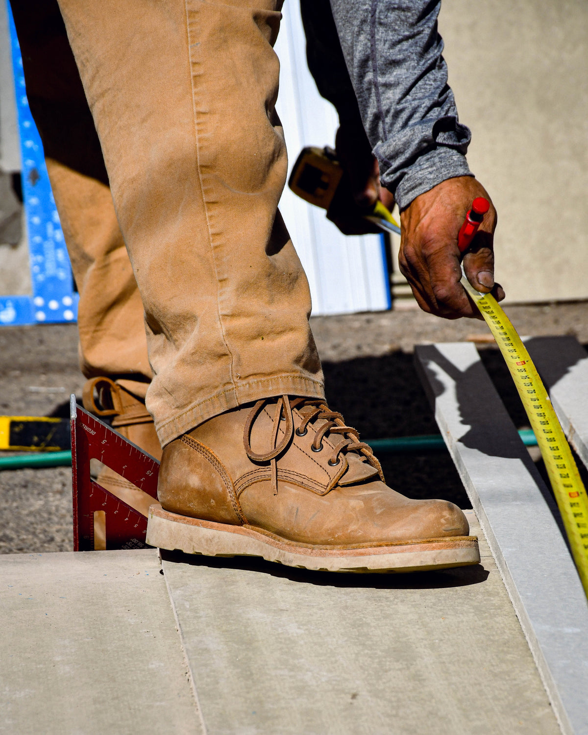Model wearing tan leather boots and measuring a plank while standing half on a platform and half on gravel.