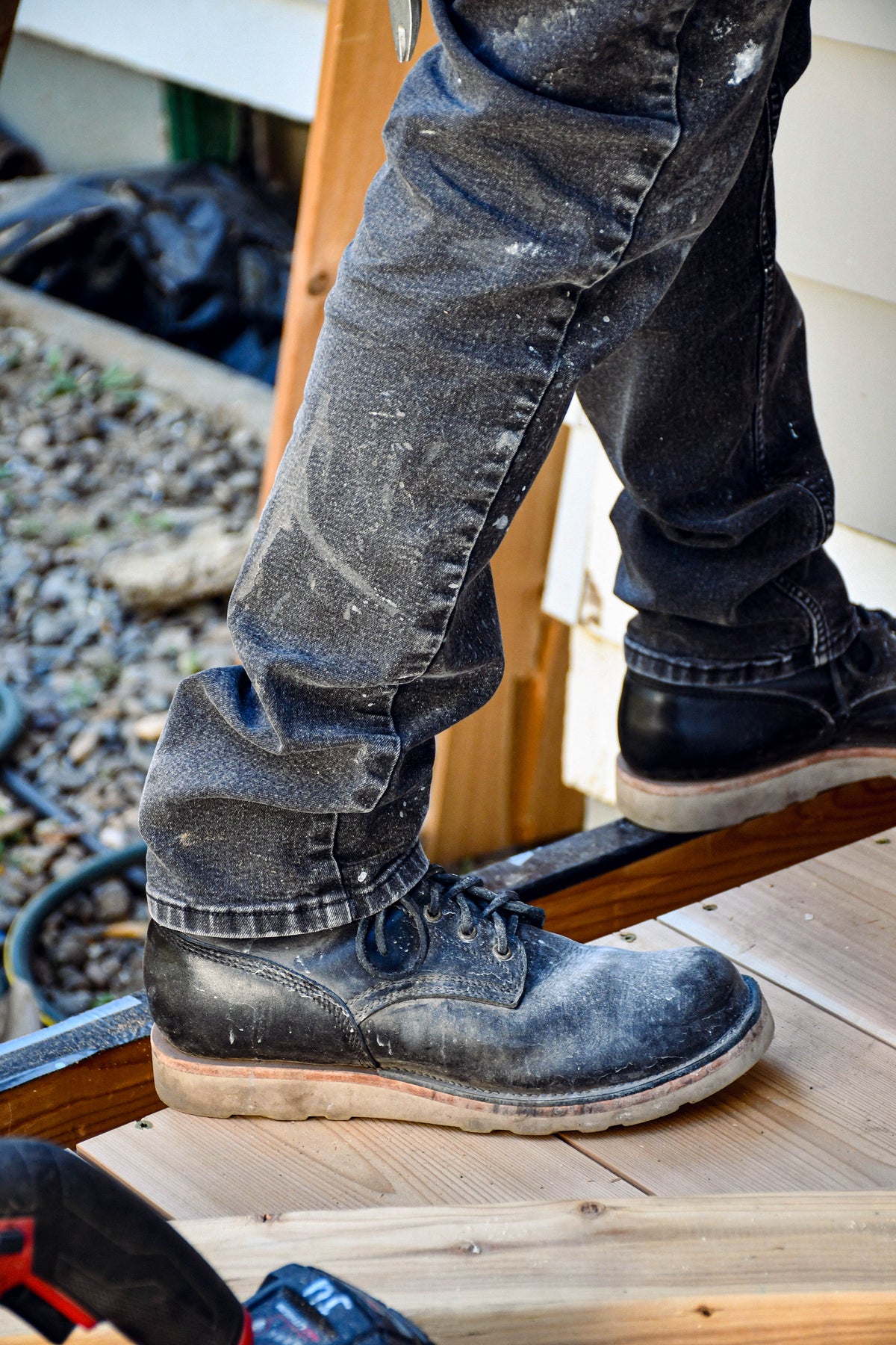 Model wearing dusty black leather boots and black jeans. Standing on unfinished deck with tools in foreground. 
